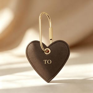Könnte beinhalten: Ein brauner herzförmiger Leder-Schlüsselanhänger mit goldfarbener Hardware. Der Schlüsselanhänger trägt den Schriftzug "TO" in goldfarbenen Buchstaben. Der Verschluss hat eine goldfarbene, längliche ovale Form.