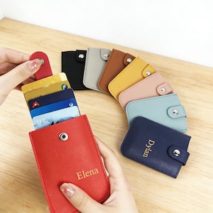 Puede incluir: Una colección de tarjeteros rectangulares en varios colores, incluyendo rojo, azul marino, azul claro, rosa, amarillo, marrón, gris y negro. Cada tarjetero tiene un cierre de botón a presión y un nombre grabado en oro, como Elena y Dylan.