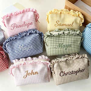 Puede incluir: Una colección de estuches rectangulares de tela con estampado de cuadros vichy en varios colores, incluyendo rosa, amarillo, azul, verde y marrón. Cada estuche tiene un borde superior con volantes, cremallera y un nombre bordado al frente.