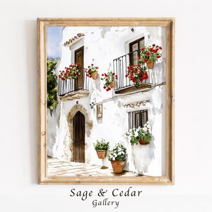 Könnte beinhalten: Ein gerahmtes Aquarellbild, das ein weißes mediterranes Gebäude mit Bogentüren, dunklen Holztüren und schwarzen Eisenbalkonen zeigt. Topfpflanzen mit leuchtend roten und weißen Blüten sind auf den Balkonen und an den Wänden angebracht.