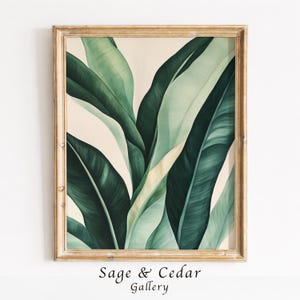Könnte beinhalten: Ein gerahmter Kunstdruck mit großen, überlappenden tropischen Bananenblättern in verschiedenen Schattierungen von tiefem Waldgrün und hellem Salbei vor einem cremefarbenen Hintergrund. Das Kunstwerk befindet sich in einem strukturierten, hellbraunen Holzrahmen.