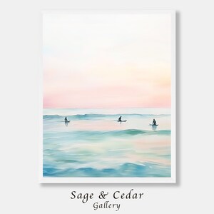 Könnte beinhalten: Ein gerahmter Kunstdruck mit einer Ozeanszene im Aquarellstil, drei Surfer auf ihren Brettern im türkisfarbenen Wasser unter einem sanften rosa-weißen Sonnenuntergangshimmel. Der Text Sage & Cedar Gallery ist unten schwarz aufgedruckt.