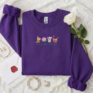 May include: A purple sweatshirt with rolled cuffs, featuring an embroidered design of a boat, flower, pig, and drink. The text "how far I'll go" is embroidered below the images. A white rose and jewelry are in the background.