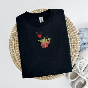 May include: Black t-shirt featuring an embroidered design of a green alien-like character holding a red cup and a Mickey Mouse balloon. The shirt is displayed on a woven basket with denim shorts and white sneakers in the background.
