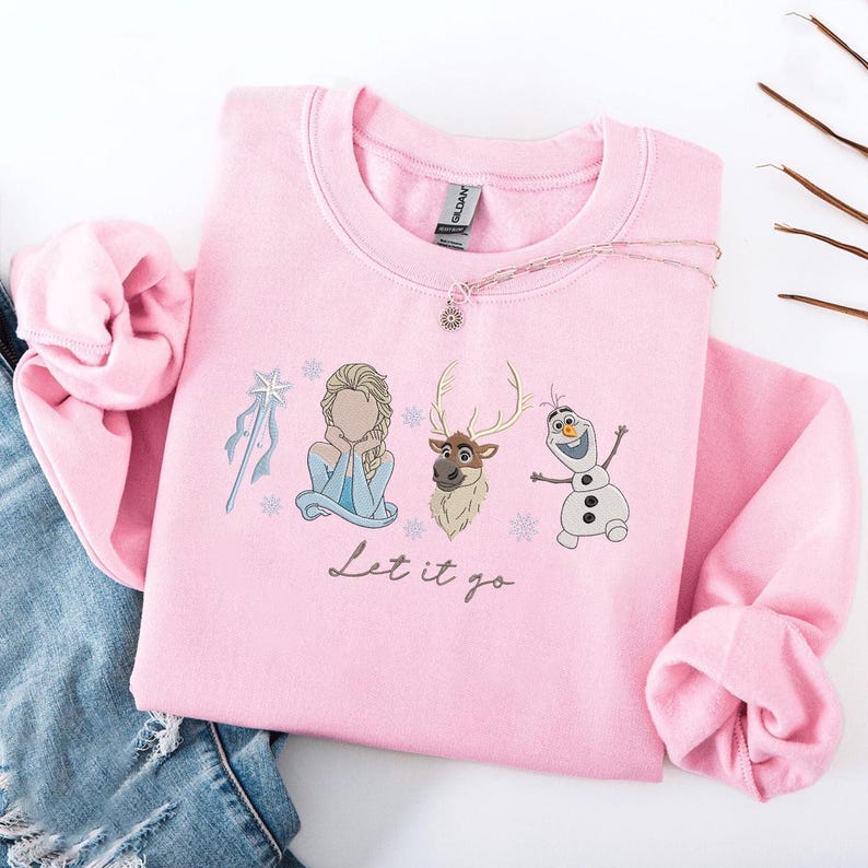May include: A light pink sweatshirt featuring a graphic of characters from the movie Frozen, including Elsa, Sven, and Olaf, with the phrase "Let it go" in script. The sweatshirt has a crew neck and long sleeves.