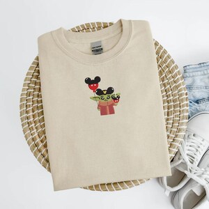 May include: A beige crew neck t-shirt featuring an embroidered design of a green character holding Mickey Mouse balloons. The character is wearing a red coat. The shirt is folded on a woven basket, with denim shorts and white sneakers in the background.