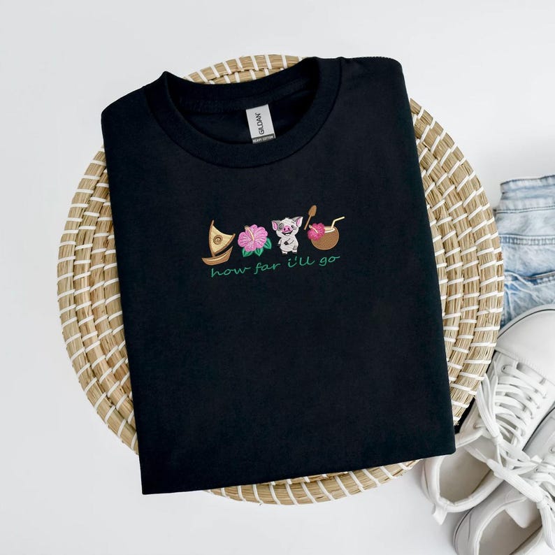 May include: Black t-shirt with embroidered design featuring a boat, flowers, a pig, and a coconut drink. The phrase "how far I'll go" is embroidered in green below the images. The shirt is displayed on a woven basket.