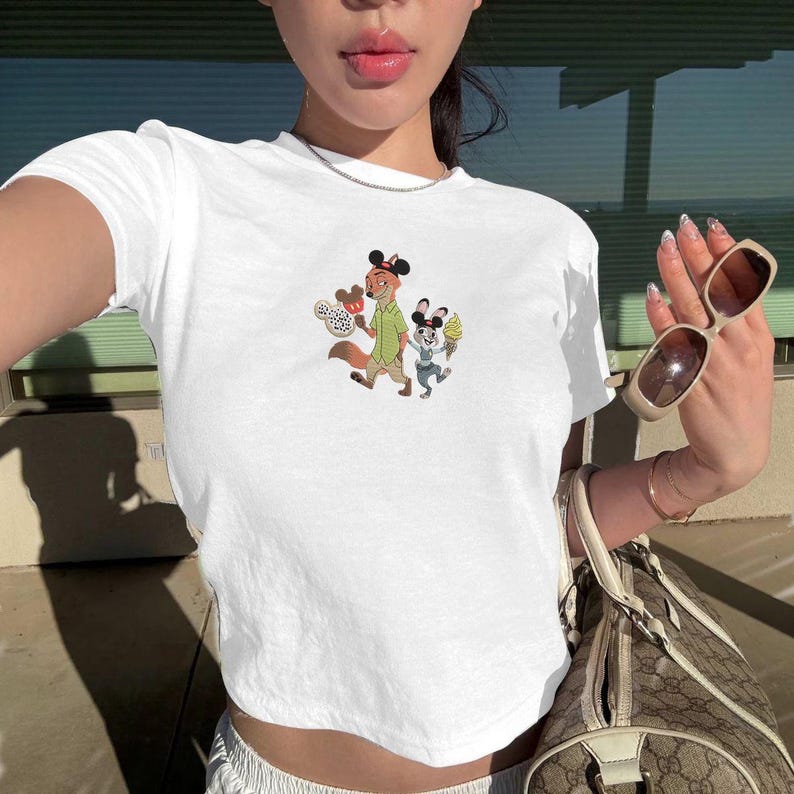 May include: White crop top featuring a colorful cartoon graphic of two characters. The graphic depicts a fox and a mouse holding treats. The shirt has short sleeves and a crew neck.