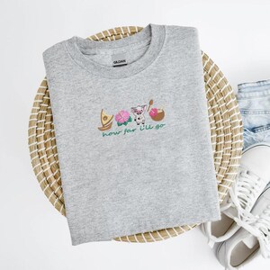 May include: A heather gray crew neck sweatshirt featuring an embroidered design with a boat, pink flowers, a pig, and a bowl. The text "how far I'll go" is below the design. The sweatshirt is made of a soft material.