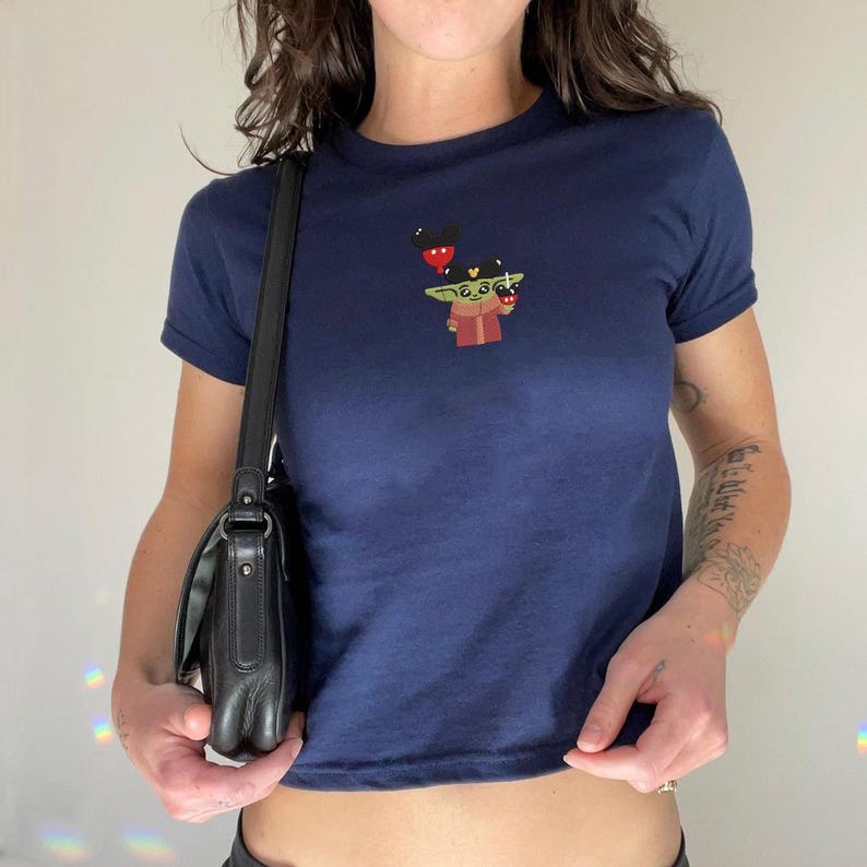 May include: Navy blue t-shirt with a small embroidered design featuring a cartoon character holding a red object and wearing a hat with Mickey Mouse ears. The shirt has short sleeves.