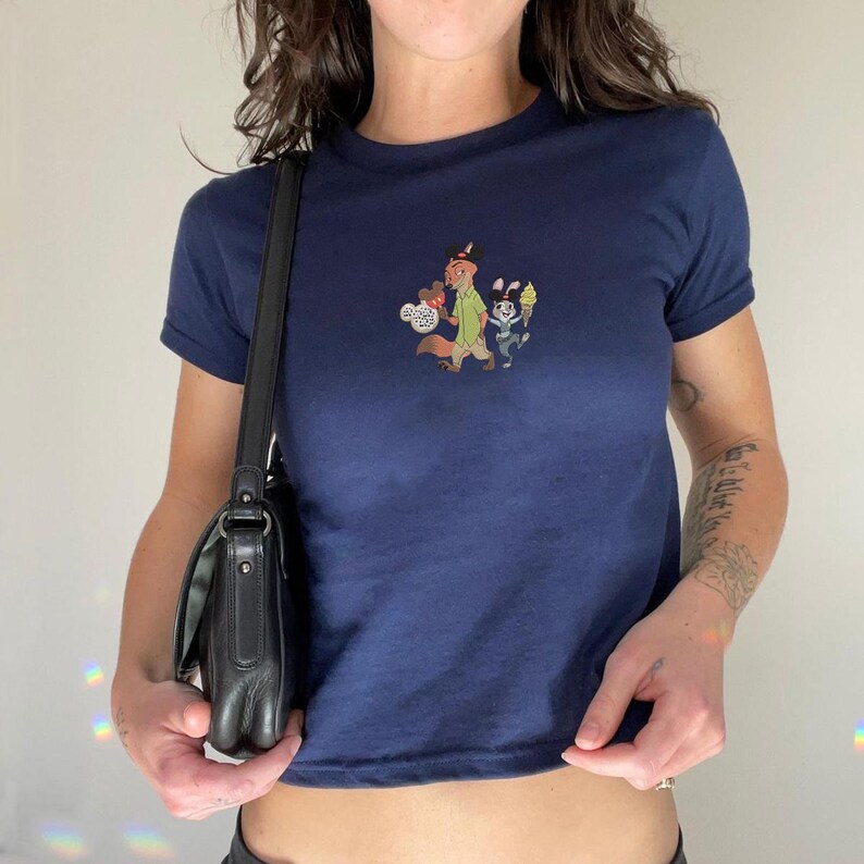 May include: Navy blue t-shirt with a crew neck and short sleeves. The shirt features a colorful graphic of cartoon characters. A black leather shoulder bag with a flap closure hangs from the shoulder.