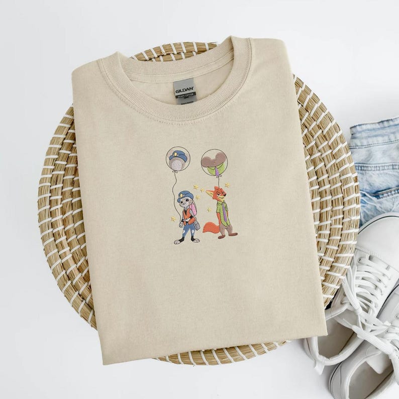 May include: A beige crewneck sweatshirt featuring an embroidered design of two cartoon characters holding balloons. One character is a rabbit in a police uniform, and the other is a fox in a green jacket. The sweatshirt is displayed on a woven basket.