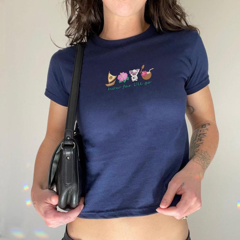 May include: Navy blue crop top with embroidered tropical designs, including a boat, flowers, and a coconut drink. The text "how far I'll go" is embroidered below the designs. The shirt has short sleeves.