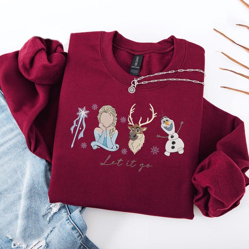 May include: A maroon crewneck sweatshirt featuring a graphic of characters from the movie Frozen, including Elsa, Olaf, and Sven, with the phrase "Let it go." A silver necklace is draped across the sweatshirt.