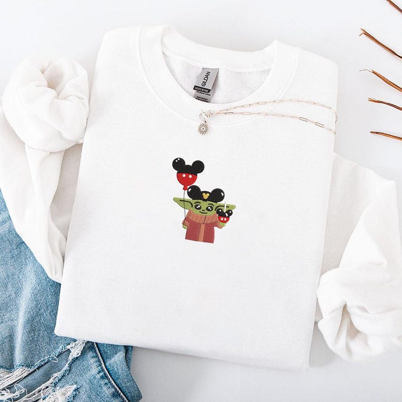 May include: White sweatshirt featuring a cartoon character holding a Mickey Mouse balloon. The character is wearing a red robe and a green scarf. A silver necklace with a small pendant is also visible.