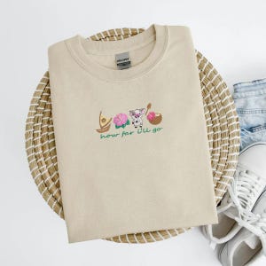 May include: Beige crew neck sweatshirt with embroidered design featuring a boat, pink flowers, a pig, and a coconut. The words "how far I'll go" are embroidered below the design. The sweatshirt is folded on a woven mat.