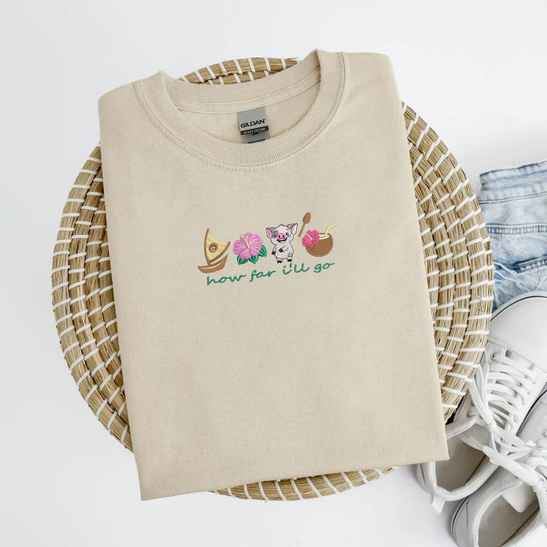 May include: Beige crew neck sweatshirt with embroidered design featuring a boat, pink flowers, a pig, and a coconut. The words "how far I'll go" are embroidered below the design. The sweatshirt is folded on a woven mat.