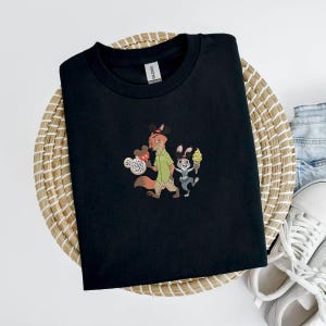 May include: Black t-shirt featuring an embroidered design of a fox and a rabbit holding ice cream cones. The fox wears a green shirt and the rabbit wears a gray outfit. The t-shirt is displayed on a woven basket.