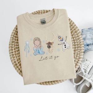 May include: Beige crewneck sweatshirt featuring embroidered images of Elsa, Sven, and Olaf from Frozen, with the phrase "Let it go" in script. The sweatshirt is folded on a woven basket.