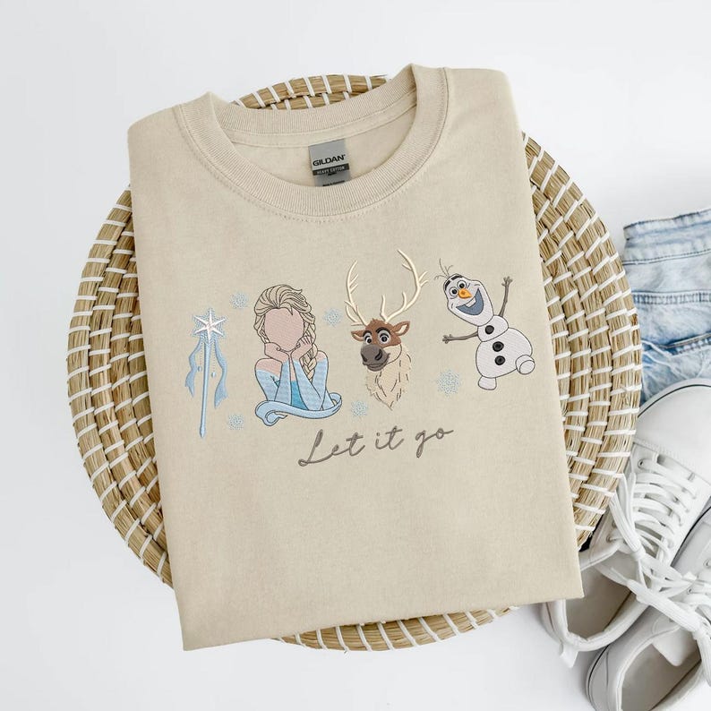 May include: Beige crewneck sweatshirt featuring embroidered images of Elsa, Sven, and Olaf from Frozen, with the phrase "Let it go" in script. The sweatshirt is folded on a woven basket.