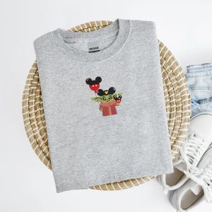 May include: A light gray crew neck sweatshirt featuring an embroidered design of a green character holding red and black Mickey Mouse balloons. The character is wearing a red and brown outfit. The sweatshirt is on a woven basket.