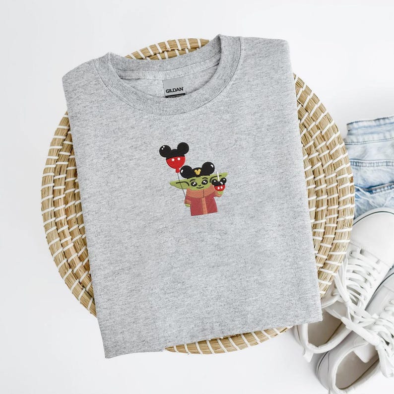 May include: A light gray crew neck sweatshirt featuring an embroidered design of a green character holding red and black Mickey Mouse balloons. The character is wearing a red and brown outfit. The sweatshirt is on a woven basket.