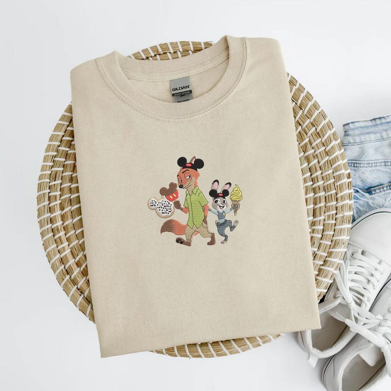 May include: Beige crew neck sweatshirt with an embroidered design featuring a fox and a rabbit, both wearing Mickey Mouse ears and holding ice cream cones. The sweatshirt is folded on a woven basket with denim and white sneakers in the background.