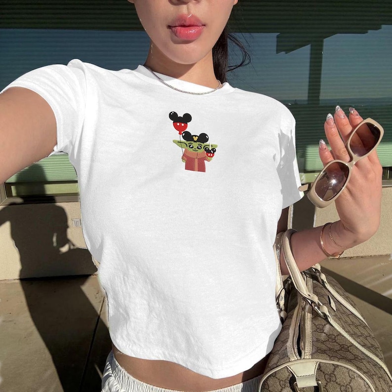 May include: White crop top featuring a cartoon graphic of a character holding a Mickey Mouse balloon. The graphic is in shades of green, red, and black. The shirt is made of a soft, lightweight material.