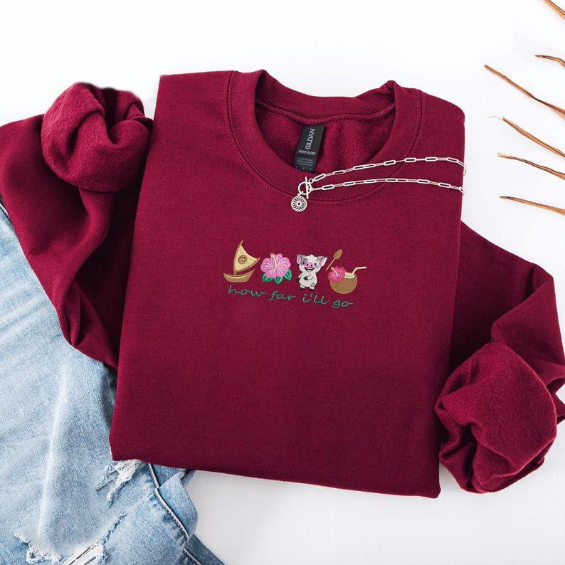 May include: A maroon crewneck sweatshirt with embroidered images of a boat, flower, pig, and drink. The text "how far I'll go" is embroidered below the images. A silver necklace with a pendant is draped on the sweatshirt.
