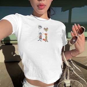 May include: White crop top featuring a cartoon illustration of two characters holding balloons. One character is a rabbit in a police uniform, and the other is a fox in a green jacket. The balloons are gray and brown.