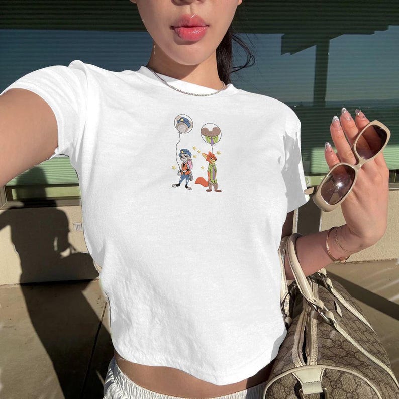 May include: White crop top featuring a cartoon illustration of two characters holding balloons. One character is a rabbit in a police uniform, and the other is a fox in a green jacket. The balloons are gray and brown.