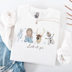 May include: White sweatshirt featuring embroidered images of characters from the movie "Frozen", including Elsa, Olaf, and Sven, along with snowflakes and a wand. The phrase "Let it go" is written in a script font below the characters. A silver necklace with a pendant is draped on the sweatshirt.