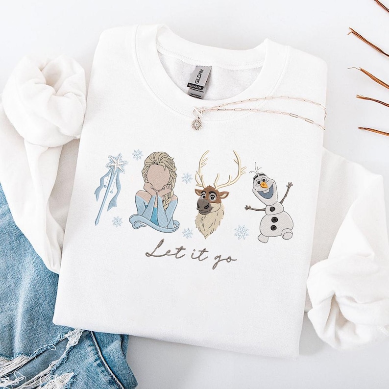 May include: White sweatshirt featuring embroidered images of characters from the movie "Frozen", including Elsa, Olaf, and Sven, along with snowflakes and a wand. The phrase "Let it go" is written in a script font below the characters. A silver necklace with a pendant is draped on the sweatshirt.