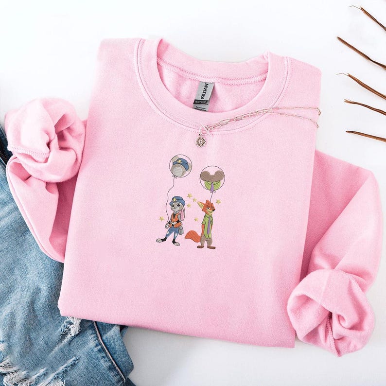 May include: A light pink sweatshirt featuring an embroidered design of two cartoon characters holding balloons. One character is a rabbit wearing a police uniform, and the other is a fox in a green shirt. A silver necklace with a sun pendant is on the sweatshirt.