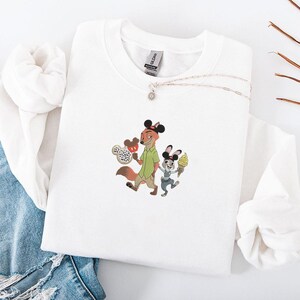 May include: White sweatshirt featuring an embroidered design of a fox and a rabbit, each wearing Mickey Mouse ears and holding ice cream cones. The fox is wearing a green shirt. A silver necklace with a small pendant is also visible.