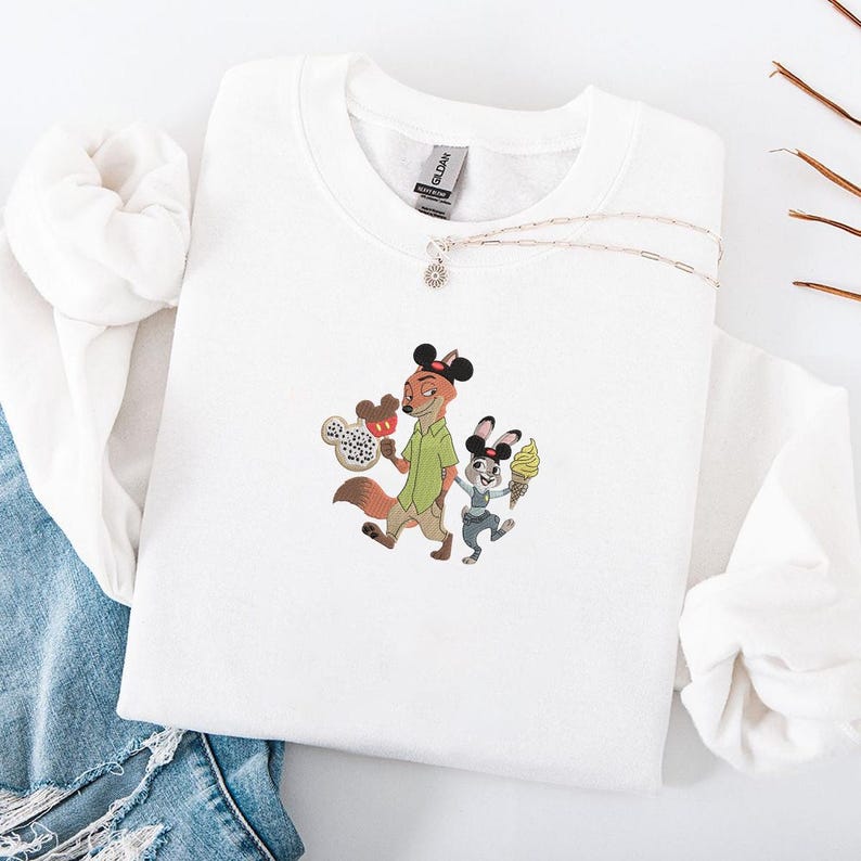 May include: White sweatshirt featuring an embroidered design of a fox and a rabbit, each wearing Mickey Mouse ears and holding ice cream cones. The fox is wearing a green shirt. A silver necklace with a small pendant is also visible.