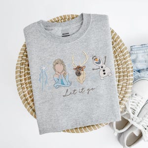 May include: A light gray crew neck sweatshirt featuring embroidered images of Elsa, Sven, and Olaf from the movie Frozen. The phrase "Let it go" is written in cursive below the characters. The sweatshirt is made of a soft material.