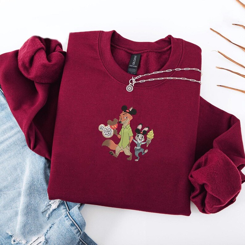 May include: A maroon sweatshirt featuring an embroidered design of two cartoon characters, one a fox and the other a rabbit, each holding ice cream cones. A silver necklace with a pendant is draped on the sweatshirt. The sweatshirt is folded on top of a pair of blue jeans.