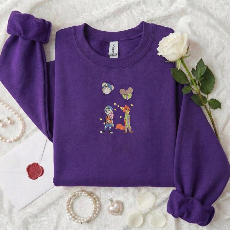 May include: A purple sweatshirt with embroidered cartoon characters. The design features a bunny, a fox, and two other small cartoon images. The sweatshirt has long sleeves with rolled cuffs. A white rose and jewelry are also in the image.