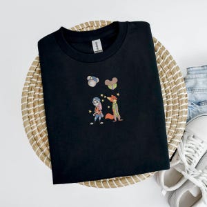 May include: Black crew neck t-shirt with embroidered cartoon characters. The design features a bunny and fox with stars, and two small character icons. The shirt is displayed on a woven basket with jeans and white sneakers.