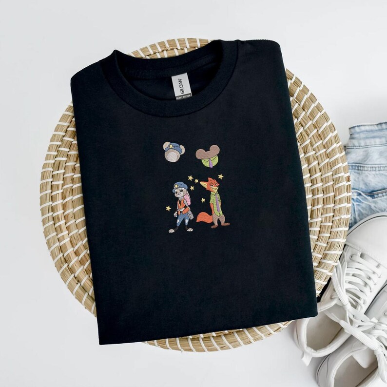 May include: Black crew neck t-shirt with embroidered cartoon characters. The design features a bunny and fox with stars, and two small character icons. The shirt is displayed on a woven basket with jeans and white sneakers.