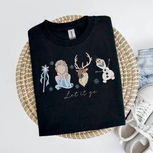 May include: Black crew neck t-shirt featuring an embroidered design with characters from the movie Frozen. The design includes Elsa, a reindeer, and Olaf the snowman, with the phrase "Let it go" below. The shirt is displayed on a woven basket.