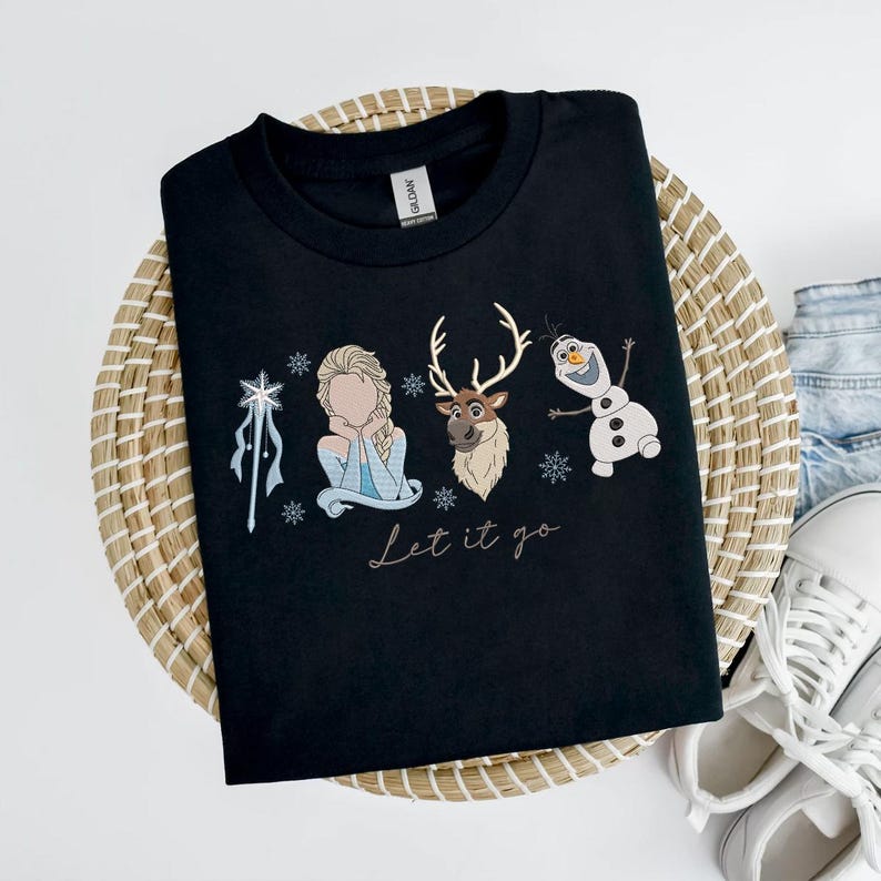 May include: Black crew neck t-shirt featuring an embroidered design with characters from the movie Frozen. The design includes Elsa, a reindeer, and Olaf the snowman, with the phrase "Let it go" below. The shirt is displayed on a woven basket.