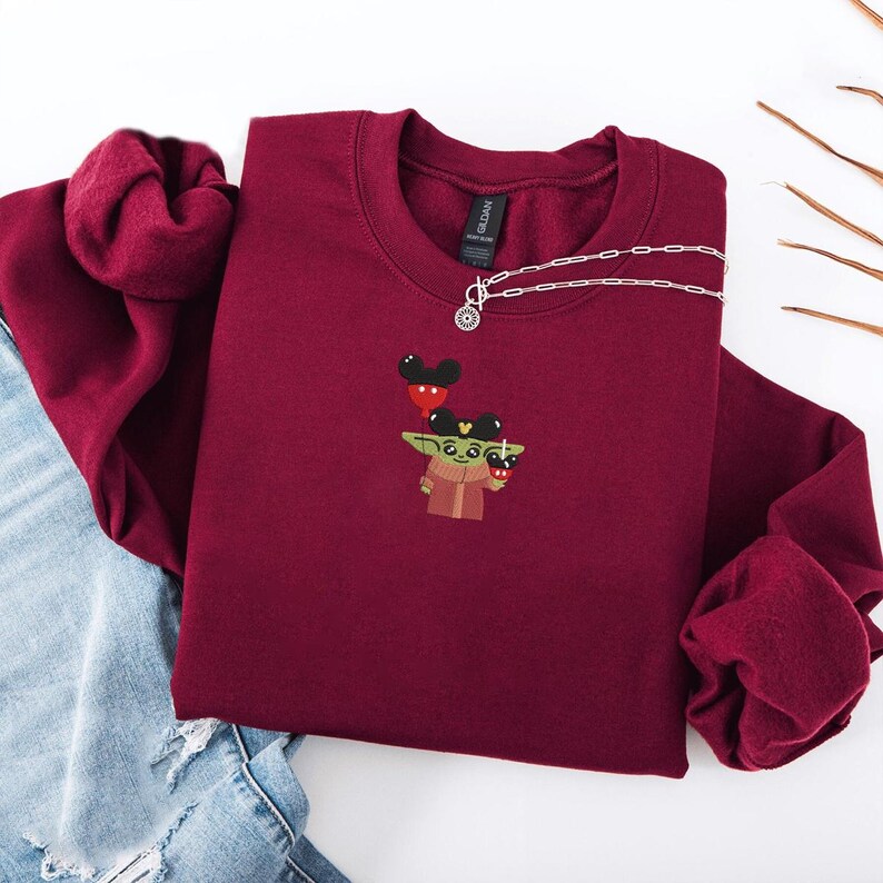 May include: A maroon crewneck sweatshirt featuring an embroidered design of a green character holding a beverage, wearing a Mickey Mouse hat. A silver necklace with a charm is draped across the sweatshirt. The sweatshirt is folded on a light blue denim surface.