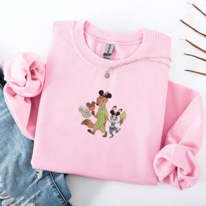 May include: Pink crewneck sweatshirt featuring an embroidered design of a fox and a rabbit holding ice cream cones and a Mickey Mouse-shaped treat. The sweatshirt has long sleeves and a round neckline. A silver necklace with a sun pendant is on the sweatshirt.