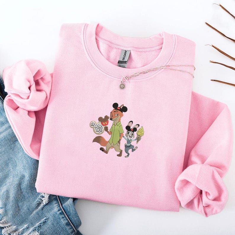 May include: Pink crewneck sweatshirt featuring an embroidered design of a fox and a rabbit holding ice cream cones and a Mickey Mouse-shaped treat. The sweatshirt has long sleeves and a round neckline. A silver necklace with a sun pendant is on the sweatshirt.