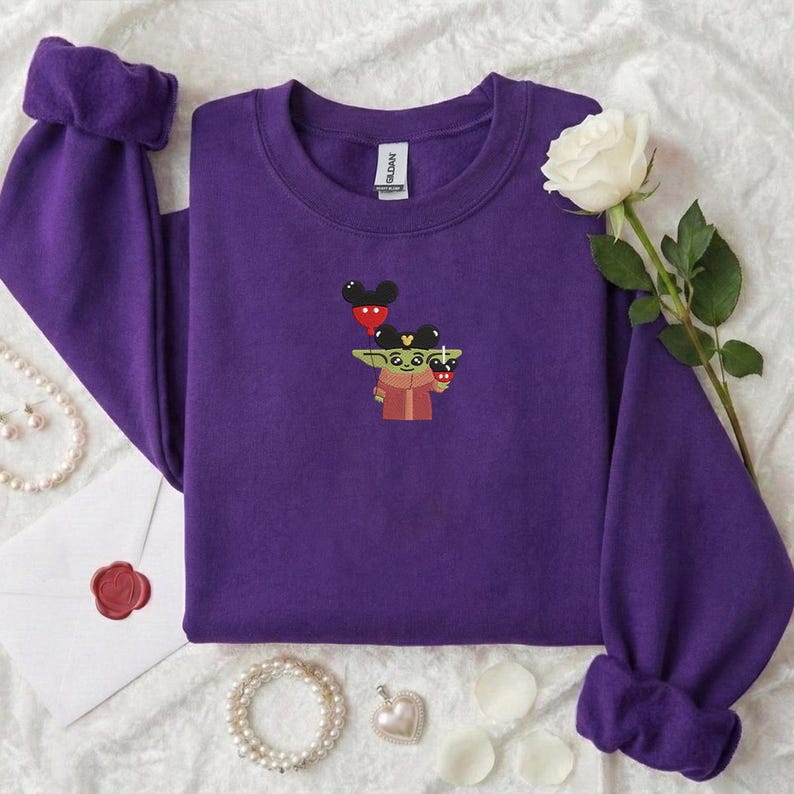 May include: A purple sweatshirt with a crew neck and long sleeves. The sweatshirt features an embroidered design of a cartoon character holding Mickey Mouse balloons and a cup. A white rose and jewelry are next to the sweatshirt.