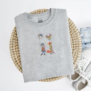 May include: A light gray crew neck sweatshirt featuring an embroidered design of two cartoon characters holding balloons. The characters are a rabbit and a fox. The sweatshirt is made of a soft material.