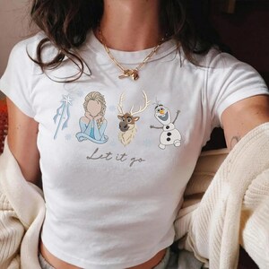 May include: White t-shirt featuring a graphic design with characters from the movie Frozen. The design includes Elsa, a reindeer, and Olaf the snowman, with the text "Let it go" in a script font. The shirt has short sleeves.