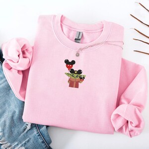 May include: A light pink sweatshirt featuring an embroidered design of a cartoon character holding a Mickey Mouse balloon. The character is wearing a red and brown outfit. A silver necklace with a sun pendant is on the sweatshirt.